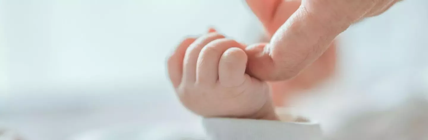 Prim-plan cu mâna unui nou-născut care ține ușor de degetul unui adult pe un fundal moale și neclar.