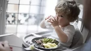 Toddler sitting in high chair by window, holding a glass of water with a plate of mixed vegetables and mashed potatoes.