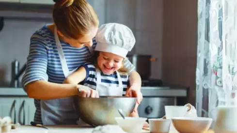 A woman helps a child in a chef hat mix ingredients in a bowl while baking in a cozy kitchen.
