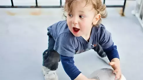 Toddler in a blue shirt crouching on the floor, holding a basketball indoors.