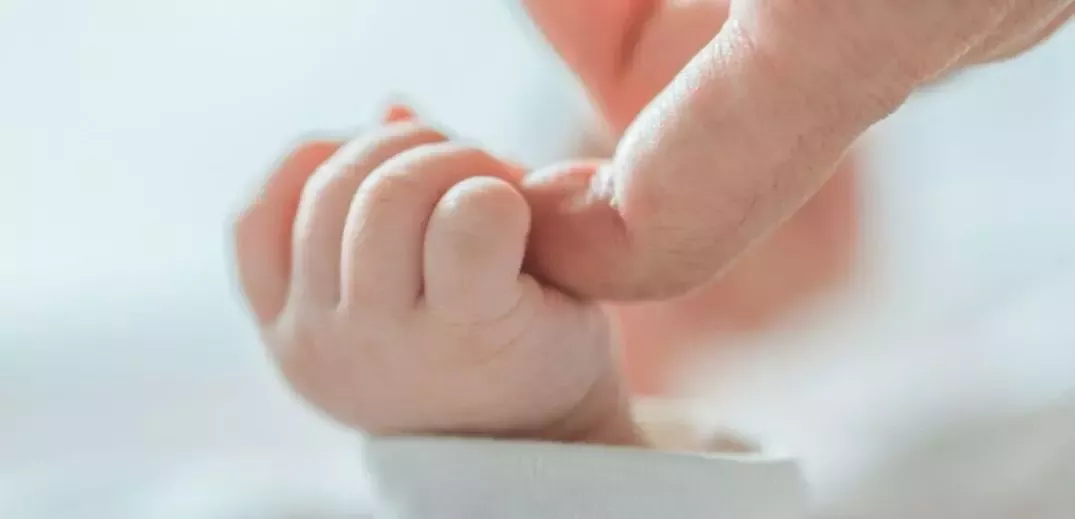 Prim-plan cu mâna unui nou-născut care ține ușor de degetul unui adult pe un fundal moale și neclar.